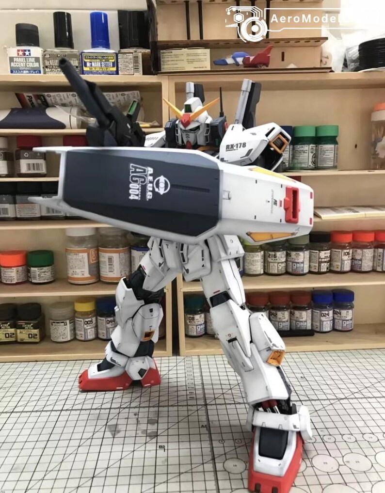 Op de afbeelding: Een gedetailleerd model van een witte, blauwe en rode robot met de tekst "AEUG AG004" op zijn schild. De robot poseert met &eacute;&eacute;n been uitgestrekt, waarbij ingewikkelde mechanische details worden getoond. Het model staat tegen een achtergrond van verfbenodigdheden.