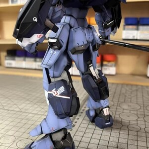 May include: A detailed, partially assembled model robot in shades of blue and black. The model features intricate mechanical details, including panels, joints, and a weapon. The AeroModelBuild logo is visible in the upper right corner. The model is on a grid surface.