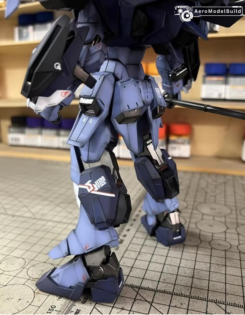 May include: A detailed, partially assembled model robot in shades of blue and black. The model features intricate mechanical details, including panels, joints, and a weapon. The AeroModelBuild logo is visible in the upper right corner. The model is on a grid surface.