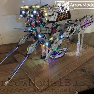 May include: A detailed model of a futuristic spacecraft, featuring a complex design with multiple sections and illuminated lights. The model is primarily white, gray, and blue, with accents of red and purple. The model is displayed on a wooden surface.
