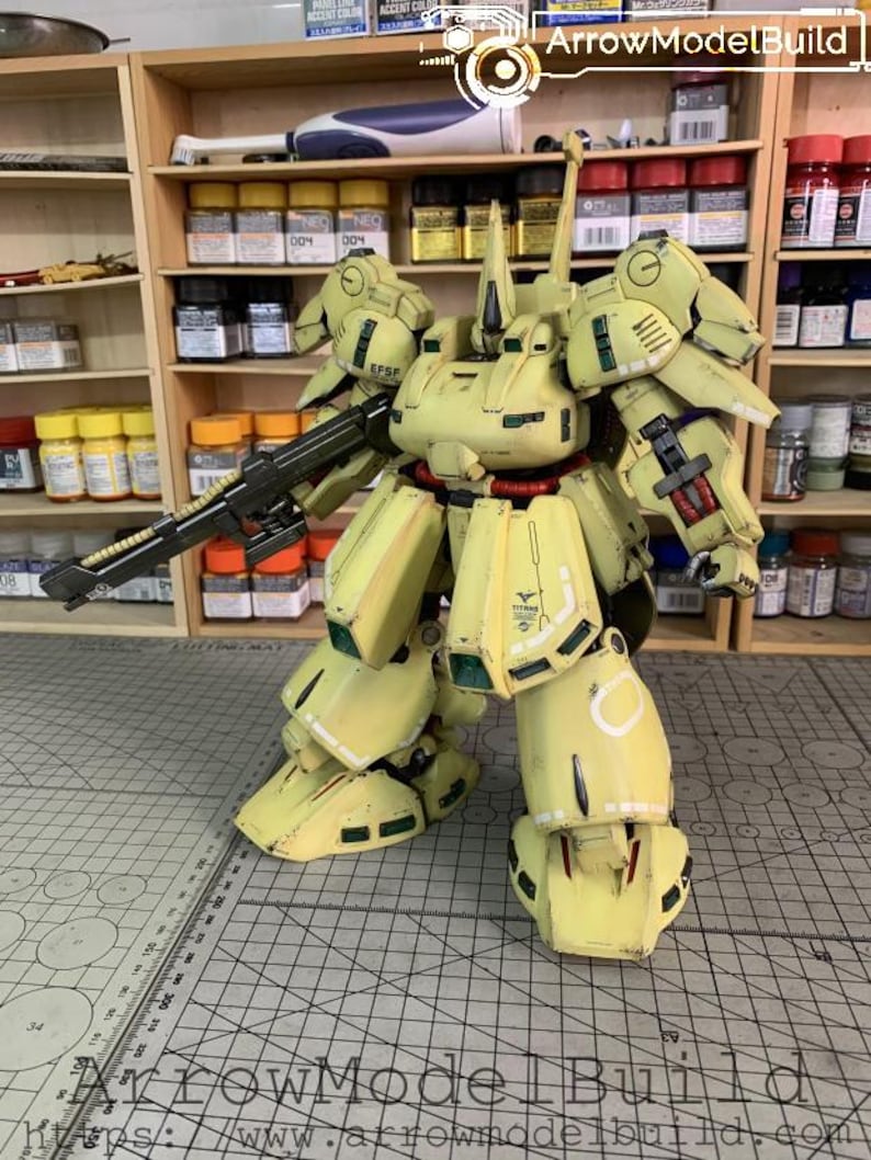 May include: A detailed, yellow model robot with a black gun, standing on a grid surface. The model has intricate details, including panels, joints, and markings. The text "EFSP" and "TITANS" are visible on the model. Shelves with paint bottles are in the background.
