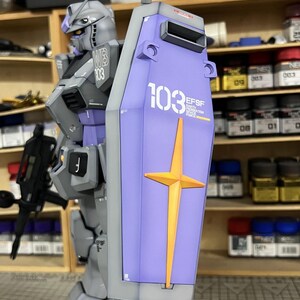 May include: A detailed model of a gray and purple robot with a shield. The shield is light purple with a gold cross and the number 103 in white. The robot has the text "WB 103" on its shoulder. The model is a Gundam.