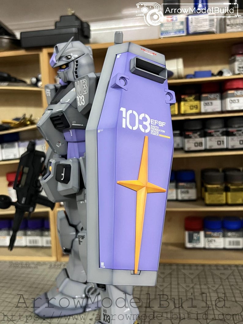 May include: A detailed model of a gray and purple robot with a shield. The shield is light purple with a gold cross and the number 103 in white. The robot has the text "WB 103" on its shoulder. The model is a Gundam.