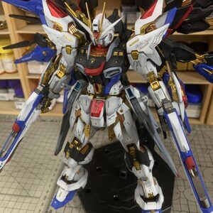 Puede incluir: Modelo detallado de un robot con detalles en blanco, azul, negro y dorado. El robot tiene grandes alas y sostiene dos armas azules y blancas. El modelo est&aacute; sobre una base negra. El texto "ArrowModelBuild" es visible.