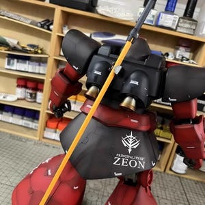 May include: A red and black model robot with a yellow pole weapon. The robot has the text "PRINCIPALITY OF ZEON" on its skirt. The model is set against a background of shelves with various tools and supplies.