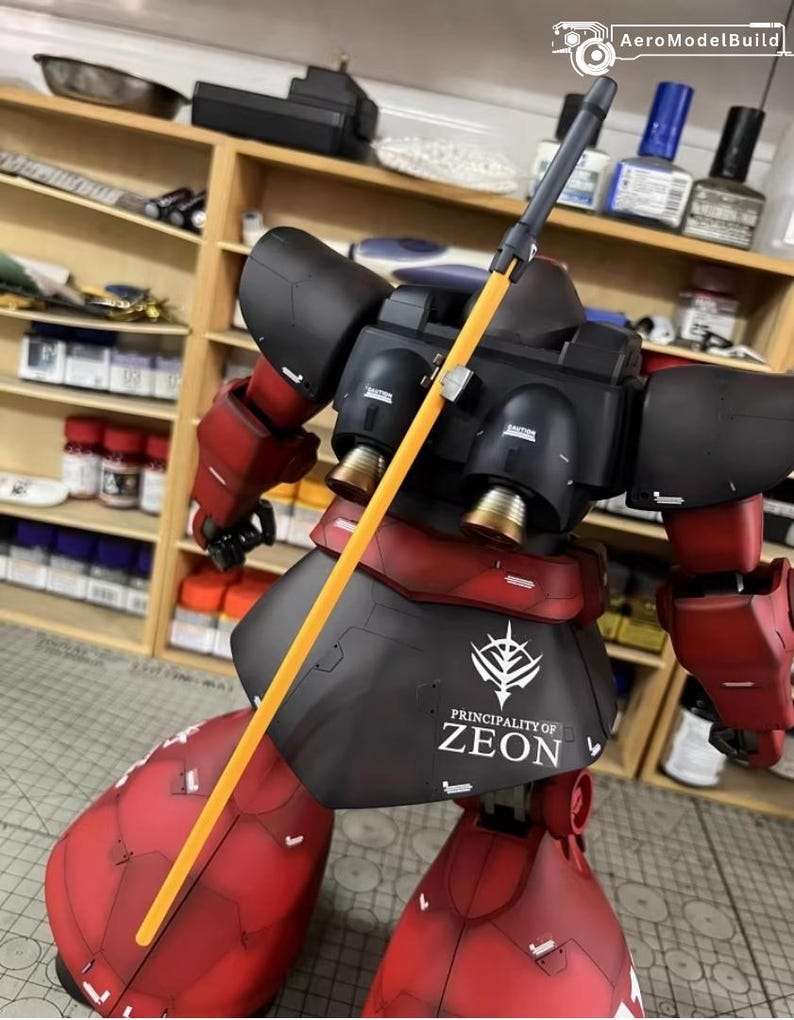 May include: A red and black model robot with a yellow pole weapon. The robot has the text "PRINCIPALITY OF ZEON" on its skirt. The model is set against a background of shelves with various tools and supplies.