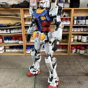 May include: A detailed model of a Gundam robot, primarily white with blue, red, yellow, and black accents. The robot is standing upright, showcasing intricate mechanical details. The model is set against a backdrop of shelves filled with paint supplies and tools.