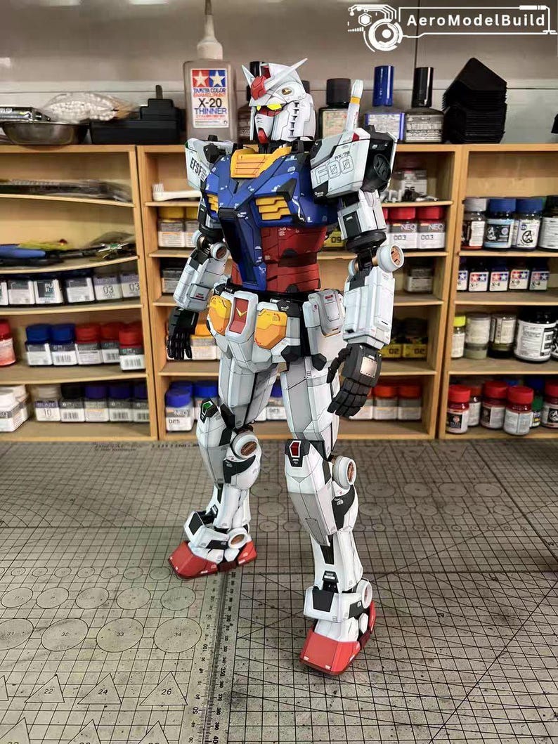 May include: A detailed model of a Gundam robot, primarily white with blue, red, yellow, and black accents. The robot is standing upright, showcasing intricate mechanical details. The model is set against a backdrop of shelves filled with paint supplies and tools.