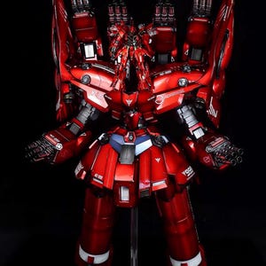 May include: A detailed, red model of a mobile suit robot with multiple weapons and armor plating. The model has a glossy finish and is displayed against a black background. The text "AeroModelBuild" is visible in the top right corner.