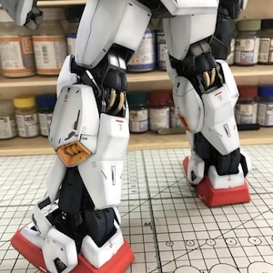 Op de afbeelding: Close-up van de benen van een gedetailleerd modelrobot. Het model heeft witte pantserplaten met zwarte en gouden accenten. De voeten zijn rood met witte details. Het model staat op een rasteroppervlak.