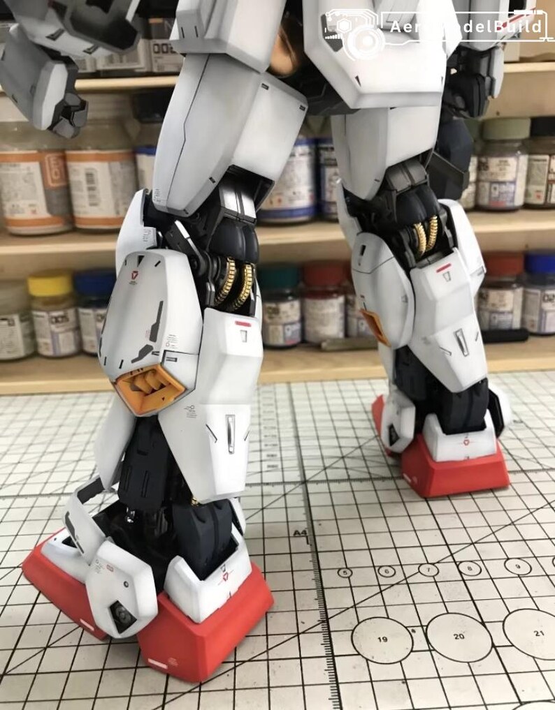 Op de afbeelding: Close-up van de benen van een gedetailleerd modelrobot. Het model heeft witte pantserplaten met zwarte en gouden accenten. De voeten zijn rood met witte details. Het model staat op een rasteroppervlak.