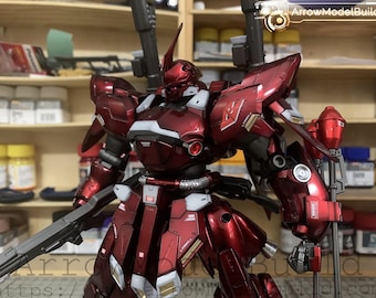 May include: A detailed, metallic red model robot with white and gold accents. The robot has a large head with a single horn, two shoulder-mounted weapons, and a large gun. The image includes the text "ArrowModelBuild".