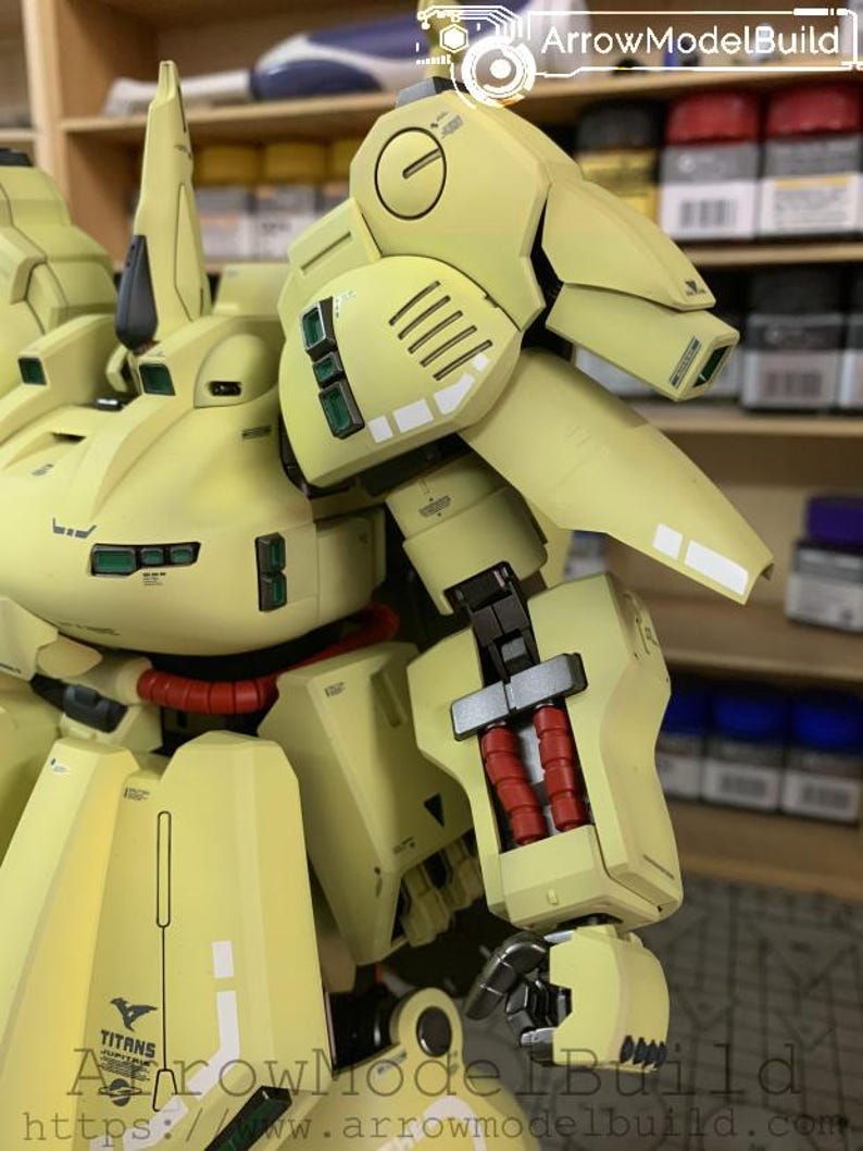 May include: A detailed, light yellow model of a robot with intricate mechanical details. The model features red cylindrical components, black accents, and the word "TITANS" printed on the lower section. The model is set against a blurred background.