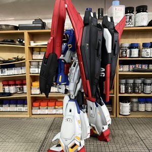 May include: A detailed model of a robot with red, black, white, and blue accents. The robot has large wings and is posed on a gray surface. The model is a complex build, showcasing intricate details and design.