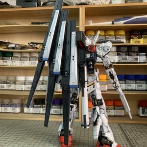 K&ouml;nnte beinhalten: Ein detailliertes Modell eines wei&szlig;en, schwarzen und roten Mecha-Roboters. Der Roboter hat gro&szlig;e Fl&uuml;gel und steht auf einer Rasteroberfl&auml;che. Der Hintergrund zeigt Regale mit Farben und Modellbauwerkzeugen.