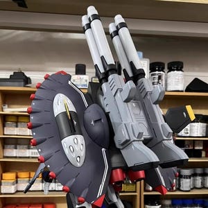 May include: A detailed model of a gray and black mech with multiple missile launchers and a large shield. The model has red and white accents, with the text "AeroModelBuild" visible in the top right corner. The model is set against a backdrop of shelves.