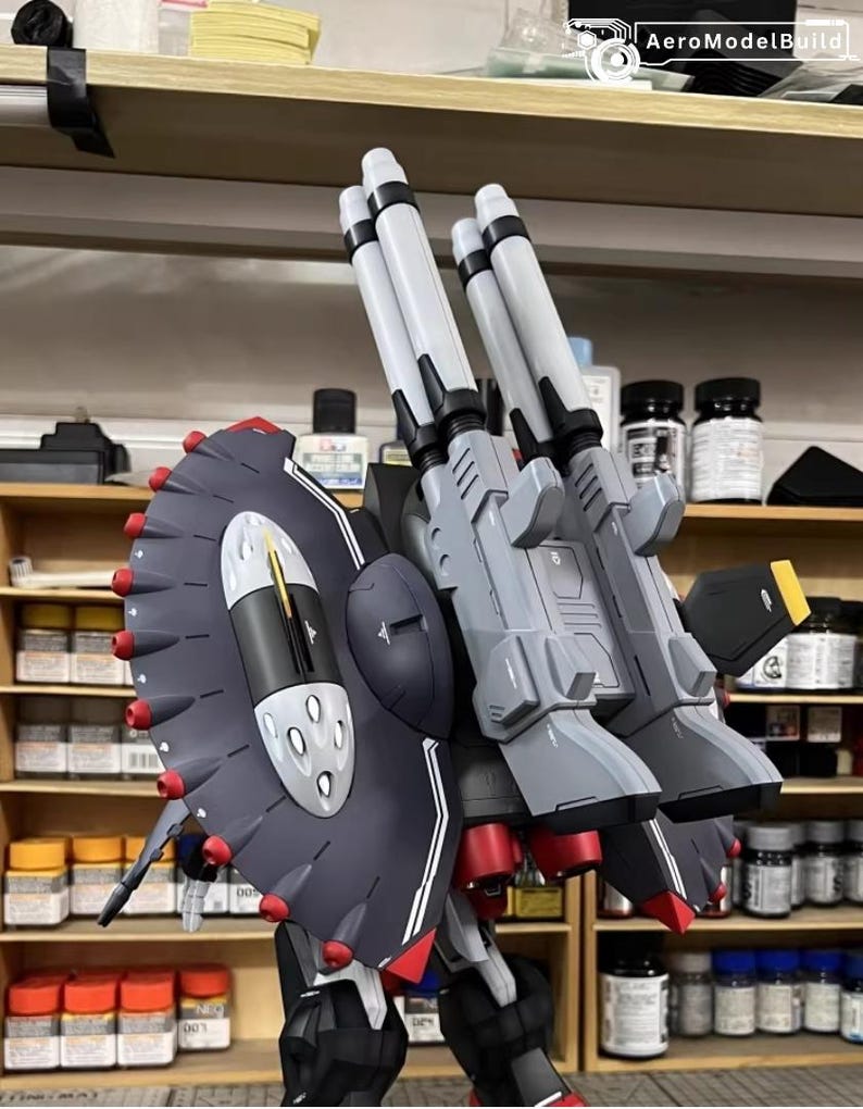 May include: A detailed model of a gray and black mech with multiple missile launchers and a large shield. The model has red and white accents, with the text "AeroModelBuild" visible in the top right corner. The model is set against a backdrop of shelves.