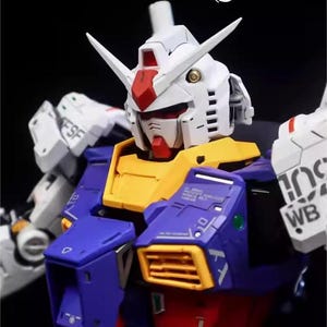 Op de afbeelding: Een close-up van een gedetailleerde, veelkleurige modelrobot. De robot heeft een witte kop met rode en gele accenten, een blauwe en gele torso en witte armen. Het model heeft ingewikkelde details en staat tegen een donkere achtergrond. De tekst "AeroModelBuild" is zichtbaar.