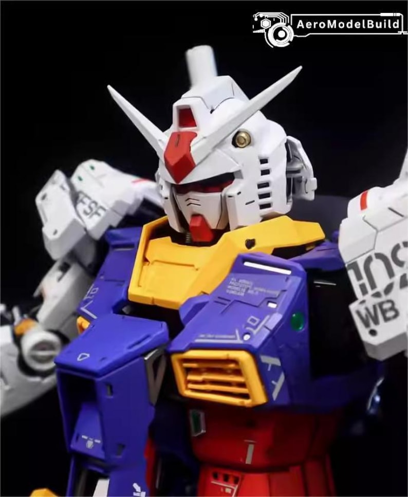 Op de afbeelding: Een close-up van een gedetailleerde, veelkleurige modelrobot. De robot heeft een witte kop met rode en gele accenten, een blauwe en gele torso en witte armen. Het model heeft ingewikkelde details en staat tegen een donkere achtergrond. De tekst "AeroModelBuild" is zichtbaar.