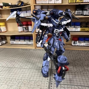 May include: A detailed, assembled model of a dark blue and gray mech robot, with multiple weapons and attachments. The model is standing on a grid-patterned surface, with shelves of paint and supplies in the background. The model has a complex design with sharp angles and intricate details.