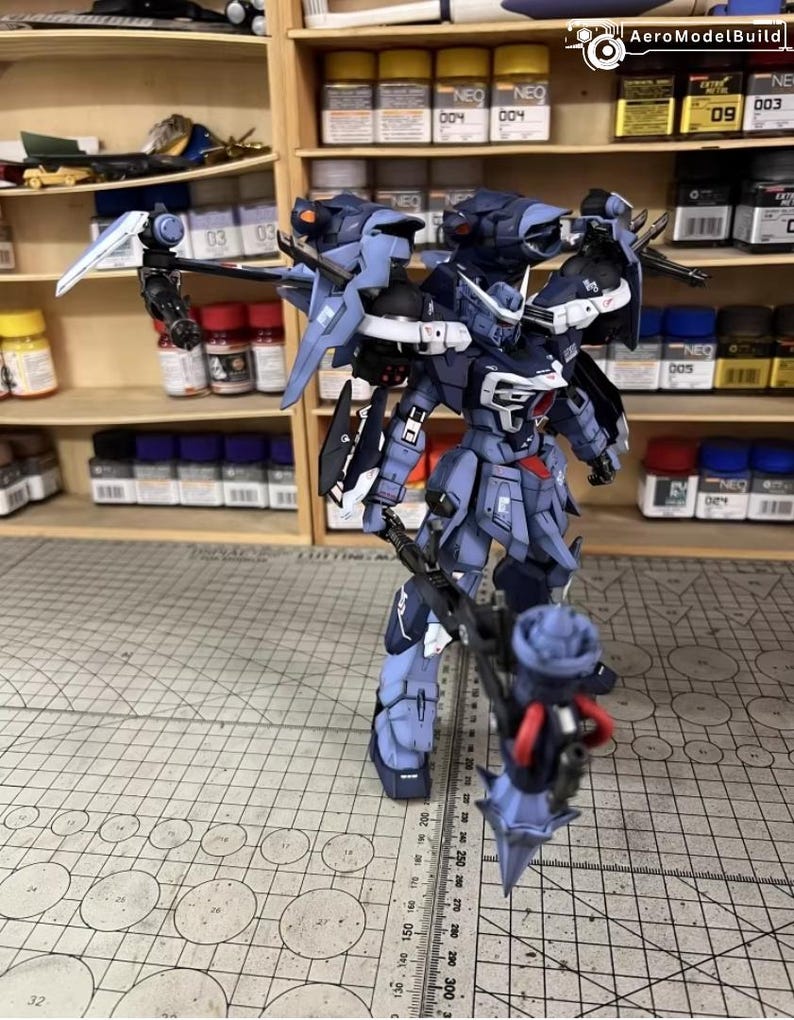 May include: A detailed, assembled model of a dark blue and gray mech robot, with multiple weapons and attachments. The model is standing on a grid-patterned surface, with shelves of paint and supplies in the background. The model has a complex design with sharp angles and intricate details.