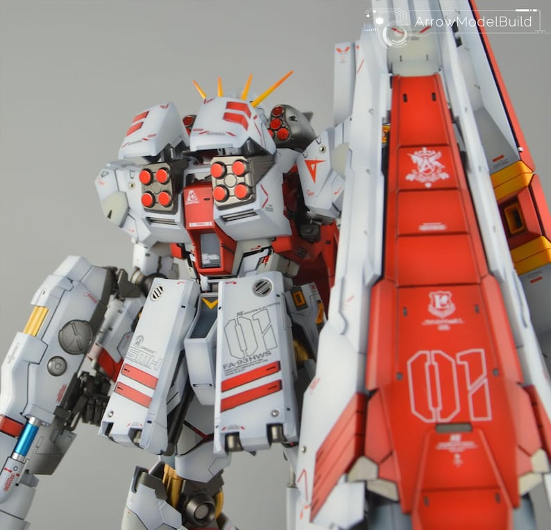 May include: A detailed model of a white and red mecha robot with intricate designs. The robot features multiple missile launchers, yellow accents, and the text "FA-93 HWS" and "01". The model is a complex assembly.