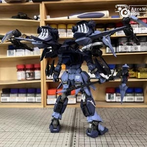 May include: A detailed model of a dark blue and black robot with multiple wings and weapons. The robot is standing on a grid surface, with shelves of paint bottles in the background. The AeroModelBuild logo is visible.