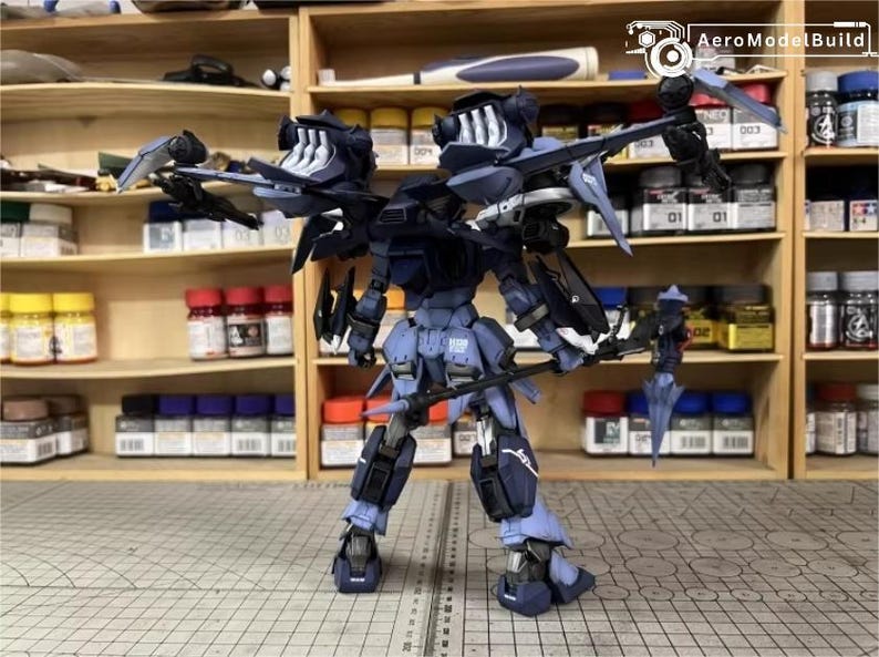 May include: A detailed model of a dark blue and black robot with multiple wings and weapons. The robot is standing on a grid surface, with shelves of paint bottles in the background. The AeroModelBuild logo is visible.