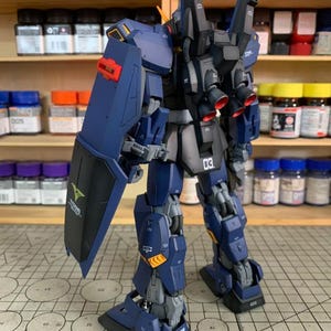 May include: A detailed model of a dark blue and black robot with red accents. The robot has a shield with a green logo and the number "03" on its leg. The model is on a patterned surface.