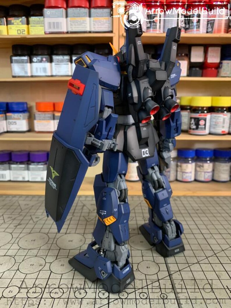 May include: A detailed model of a dark blue and black robot with red accents. The robot has a shield with a green logo and the number "03" on its leg. The model is on a patterned surface.