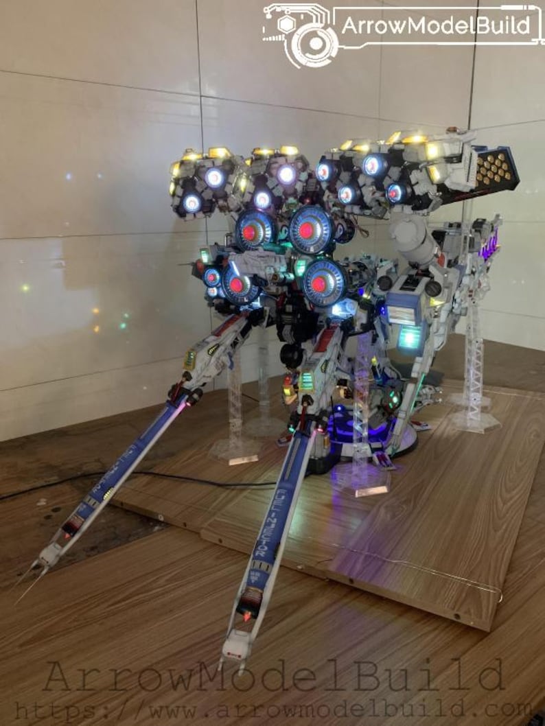 May include: A detailed model of a futuristic robot with multiple light-up components. The model features white, blue, and red accents, with the text "ArrowModelBuild" visible. The robot is displayed on a wooden surface.