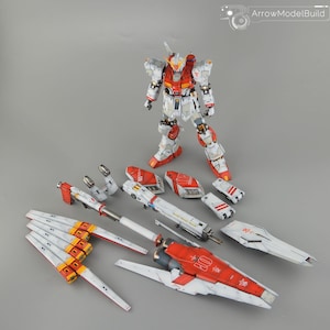 May include: A detailed model kit of a Gundam robot, featuring a white, red, and yellow colour scheme. The assembled robot stands upright, with various disassembled parts arranged around it. The kit includes wings, weapons, and armour pieces.