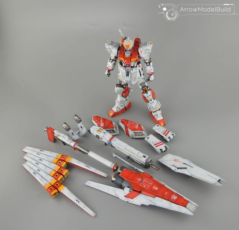 May include: A detailed model kit of a Gundam robot, featuring a white, red, and yellow colour scheme. The assembled robot stands upright, with various disassembled parts arranged around it. The kit includes wings, weapons, and armour pieces.