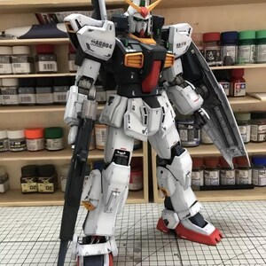 Op de afbeelding: Gedetailleerd model van een witte, zwarte en rode robot met gele en oranje accenten, die een groot wapen vasthoudt. Het model is gelabeld met RX-178 en SAG004. Het staat voor een plank met verfpotten.