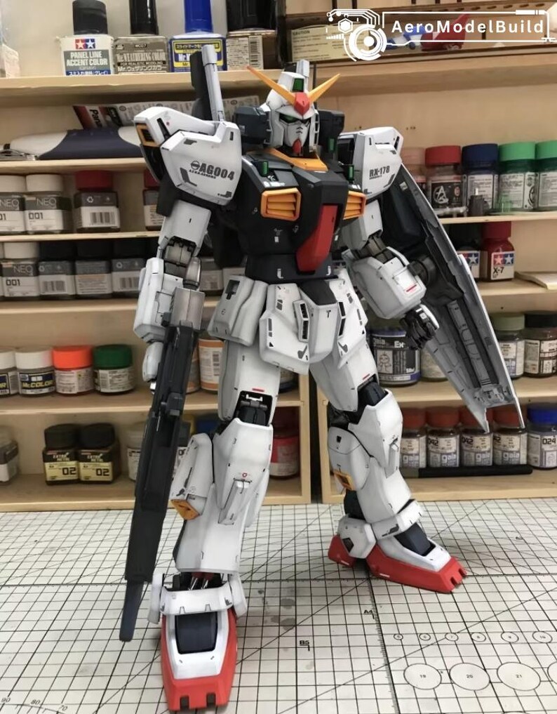Op de afbeelding: Gedetailleerd model van een witte, zwarte en rode robot met gele en oranje accenten, die een groot wapen vasthoudt. Het model is gelabeld met RX-178 en SAG004. Het staat voor een plank met verfpotten.