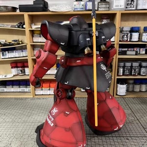 May include: A detailed model of a red and black mobile suit, featuring intricate panel lines and a yellow spear. The model has the word "ZEON" printed on the side. The model is posed on a cutting mat.