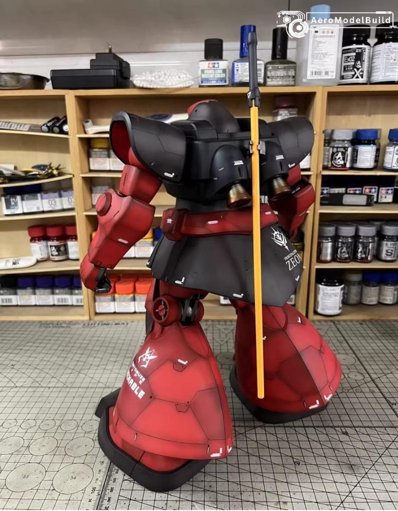 May include: A detailed model of a red and black mobile suit, featuring intricate panel lines and a yellow spear. The model has the word "ZEON" printed on the side. The model is posed on a cutting mat.