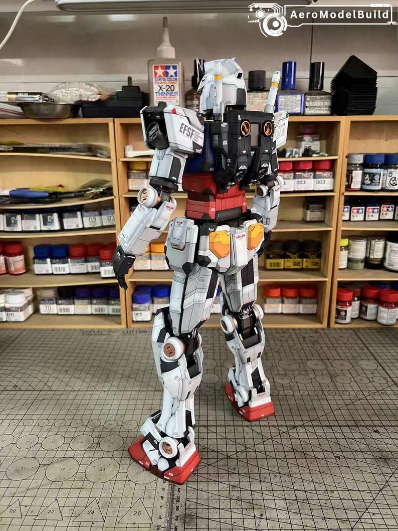 May include: A detailed, assembled Gundam model kit, primarily white with black and red accents. The model features intricate panel lines and mechanical details, with the text "EFSF" visible on the shoulder. The model is posed on a cutting mat.