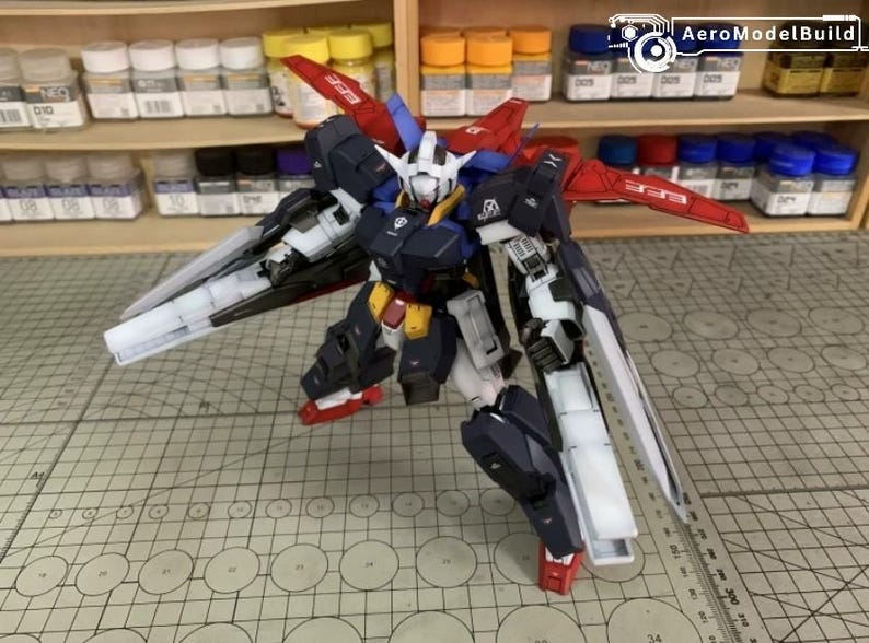 Puede incluir: Maqueta detallada de un robot mecha, con una combinaci&oacute;n de colores azul marino, blanco, rojo y amarillo. El robot tiene grandes alas y est&aacute; posado sobre una esterilla de corte. El logotipo de AeroModelBuild es visible en el fondo.