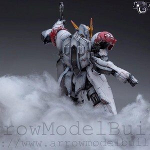 May include: A detailed model of a white and gray mech figure with red and yellow accents, posed in a dynamic stance. The figure is partially obscured by a cloud-like substance, with the text "ArrowModelBuild" visible at the bottom.