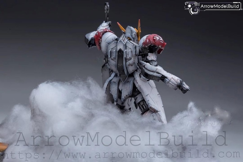 May include: A detailed model of a white and gray mech figure with red and yellow accents, posed in a dynamic stance. The figure is partially obscured by a cloud-like substance, with the text "ArrowModelBuild" visible at the bottom.