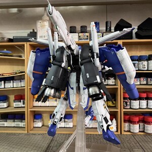 May include: A detailed model kit of a futuristic robot, with white, blue, and black components. The model is mounted on a clear stand, showcasing its intricate design and mechanical details. The background includes shelves with paint bottles and model-building supplies.