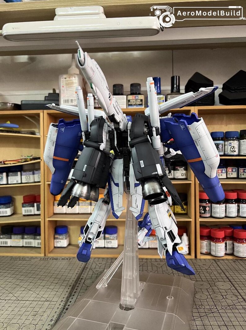 May include: A detailed model kit of a futuristic robot, with white, blue, and black components. The model is mounted on a clear stand, showcasing its intricate design and mechanical details. The background includes shelves with paint bottles and model-building supplies.
