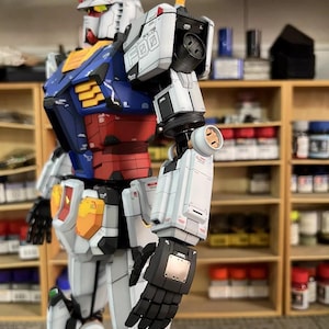 May include: A detailed model of a Gundam robot, featuring a white, blue, red, and yellow color scheme. The robot has intricate mechanical details, including segmented armor, articulated limbs, and a distinctive head with a V-fin. The model is posed in a dynamic stance.