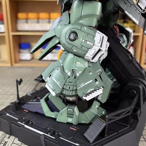 May include: A detailed, assembled model kit of a green and white mech robot. The model is on a black base with intricate panel lines and mechanical details. The image shows the robot's leg and lower torso.