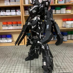 Puede incluir: Un modelo de robot negro y plateado con un dise&ntilde;o detallado, de pie sobre una superficie con patr&oacute;n de cuadr&iacute;cula. El robot tiene m&uacute;ltiples partes articuladas y est&aacute; posando con un arma. El fondo incluye estantes con varios botes de pintura.