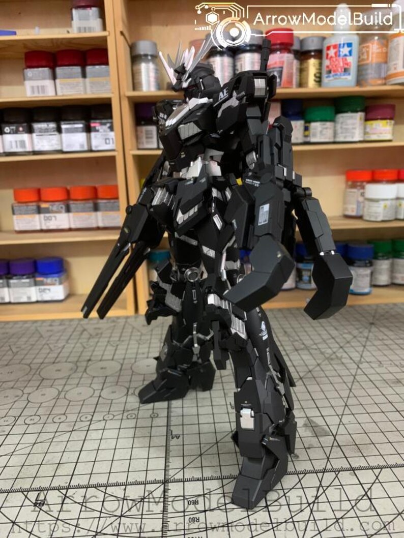 Puede incluir: Un modelo de robot negro y plateado con un dise&ntilde;o detallado, de pie sobre una superficie con patr&oacute;n de cuadr&iacute;cula. El robot tiene m&uacute;ltiples partes articuladas y est&aacute; posando con un arma. El fondo incluye estantes con varios botes de pintura.