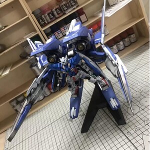 May include: A detailed model of a blue and white mecha robot, featuring intricate mechanical details and a dynamic pose. The model is mounted on a black stand, showcasing its wings and weaponry. The background includes shelves with paint bottles.