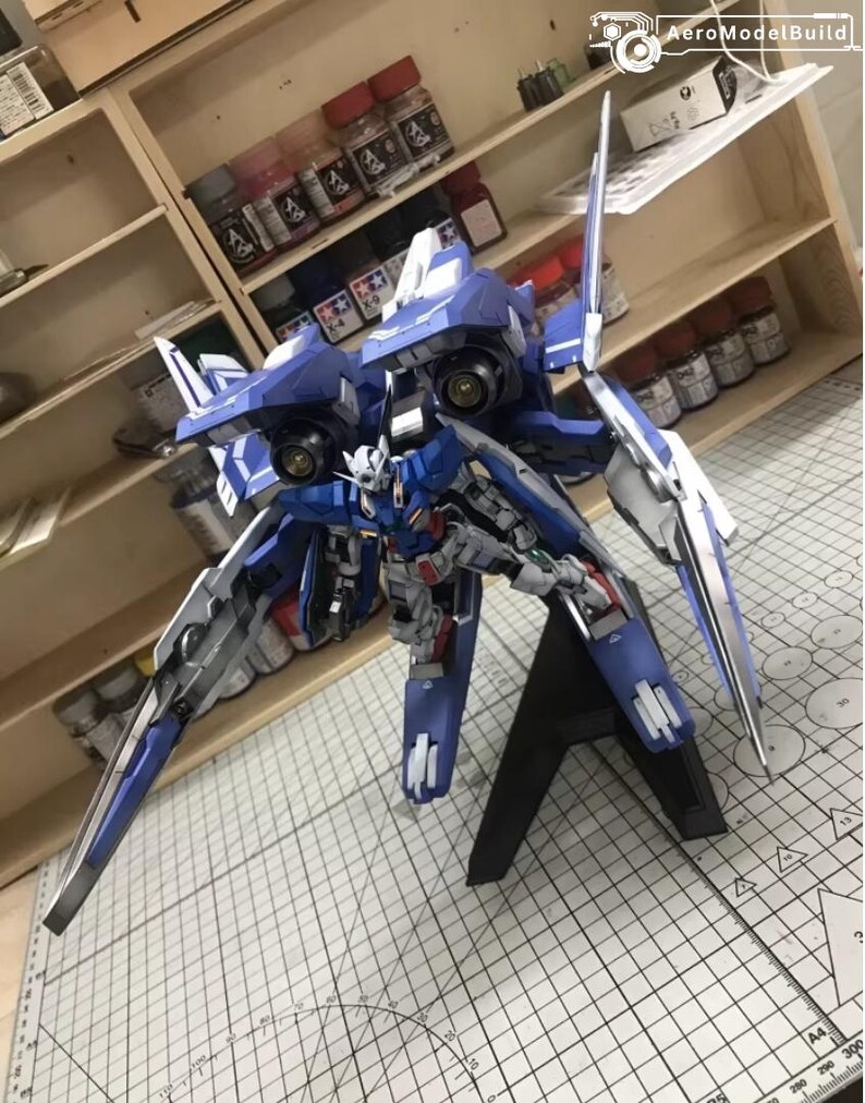 May include: A detailed model of a blue and white mecha robot, featuring intricate mechanical details and a dynamic pose. The model is mounted on a black stand, showcasing its wings and weaponry. The background includes shelves with paint bottles.