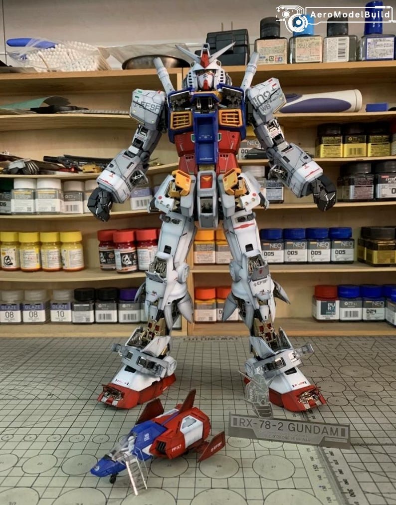 K&ouml;nnte beinhalten: Ein detailliertes Modell des RX-78-2 Gundam, ein wei&szlig;er, roter, blauer und gelber Mecha-Roboter, der aufrecht steht. Ein kleineres rotes und blaues Raumschiffmodell befindet sich davor. Das Modell steht auf einer Schneidematte mit einem Lineal.